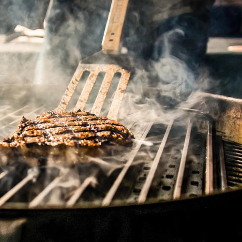 Grilled steak on a barbecue with a spatula, surrounded by smoke.